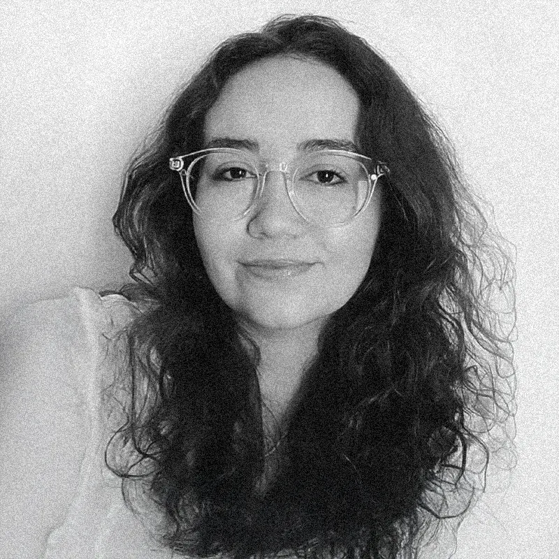 Retrato fotográfico em preto e branco de uma mulher, apresentado dentro de uma moldura circular. Ela tem a pele clara, cabelos escuros, longos e cacheados que caem sobre os ombros. Ela olha para a câmera com uma expressão serena e usa óculos de grau com armação clara e transparente. A imagem apresenta uma textura granulada, semelhante a uma técnica de impressão artística ou estêncil. O retrato circular está centralizado sobre um fundo branco com texturas sutis que lembram papel. Ela veste uma blusa clara que se confunde levemente com o fundo da foto.