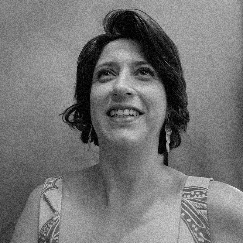 Retrato fotográfico em preto e branco de uma mulher, apresentado dentro de uma moldura circular. Ela é branca, tem cabelos escuros curtos e ondulados, e está com o rosto levemente voltado para cima, sorrindo e olhando para o alto. A mulher usa brincos compridos com três elementos circulares e uma roupa com alças largas estampadas com padrões geométricos. A imagem possui uma textura granulada, semelhante a uma impressão em papel jornal ou estêncil. O fundo é neutro e o círculo está sobreposto a uma base branca com texturas sutis.