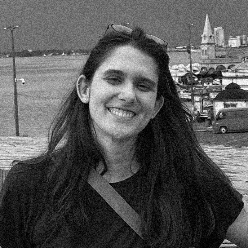 Retrato fotográfico em preto e branco de uma mulher, apresentado dentro de uma moldura circular. Ela é branca, tem cabelos escuros e longos, e sorri para a câmera. Usa óculos de sol repousados sobre a cabeça e uma camiseta escura com uma alça de bolsa atravessada pelo peito. Ao fundo, a paisagem externa mostra um corpo d'água à esquerda e uma área urbana ou portuária à direita, com construções e um veículo. Assim como nas imagens anteriores, a foto possui uma textura granulada característica de impressões em papel jornal ou técnica de estêncil, e o círculo está centralizado sobre um fundo branco texturizado.