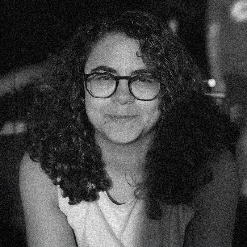 Esta é uma imagem fotográfica de retrato circular em preto e branco de uma jovem com cabelos escuros e cacheados e óculos. I imagem granulada que imita a textura do papel. O rosto da mulher está no centro do círculo, olhando diretamente para a câmera. Seus cabelos cacheados emolduram seu rosto em ambos os lados e cobrem parcialmente seus ombros. Ela está usando óculos de aro escuro e uma blusa de cor clara, possivelmente uma regata. Seu lábio superior esquerdo tem uma pequena cicatriz. A imagem está granulada e as cores são monocromáticas, criando uma sensação de antiguidade. O fundo atrás dela é escuro e desfocado, sugerindo um ambiente noturno.