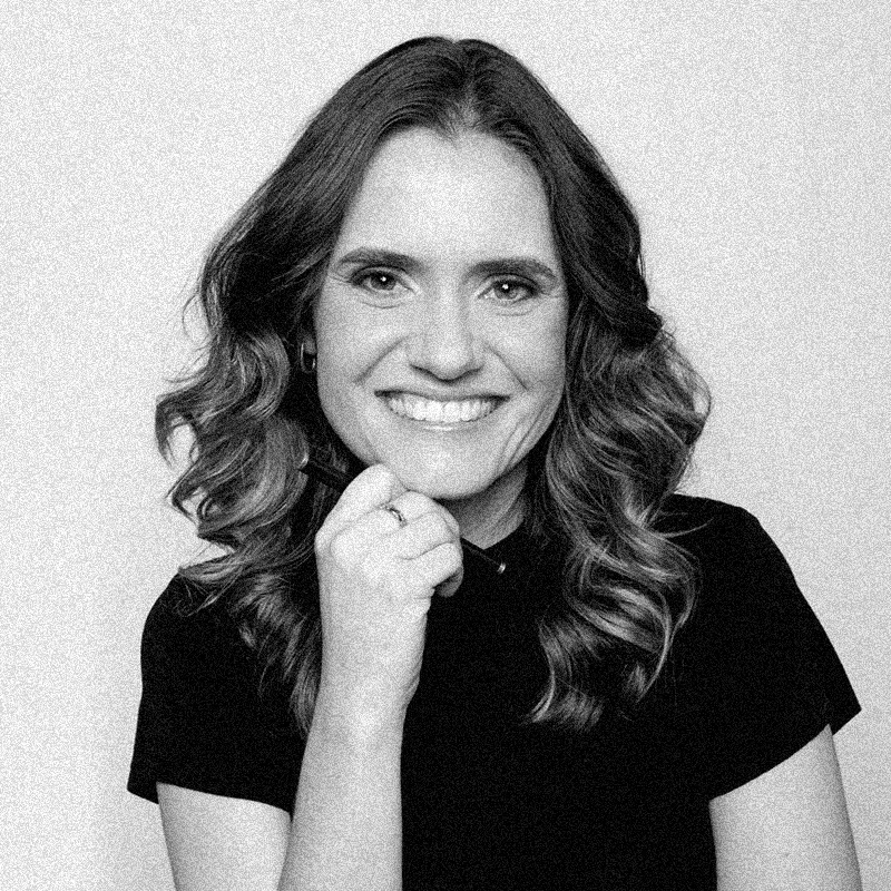 etrato fotográfico em preto e branco de uma mulher sorridente, apresentado dentro de uma moldura circular. Ela tem a pele clara e cabelos claros, longos e ondulados que caem sobre os ombros. Ela posa com a mão próxima ao queixo, segurando o que parece ser uma caneta ou pincel. Veste uma blusa escura e olha diretamente para a câmera. Assim como os demais retratos da série, a imagem possui uma textura granulada característica de impressões artísticas, e o círculo está centralizado sobre um fundo branco com texturas sutis que lembram papel.