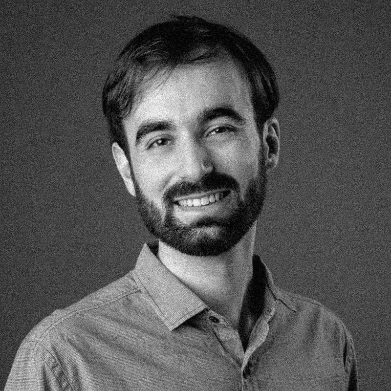 Retrato fotográfico em preto e branco de um homem jovem, apresentado dentro de uma moldura circular. Ele tem a pele branca, cabelos escuros e curtos, e uma barba rala com bigode. Ele olha diretamente para a câmera com uma expressão serena. Veste uma camiseta escura de gola redonda. O fundo do retrato é neutro e desfocado. A imagem segue a identidade visual da série, com uma textura granulada que remete a impressões em papel jornal ou estêncil. O círculo está centralizado sobre um fundo branco com texturas sutis que lembram papel.