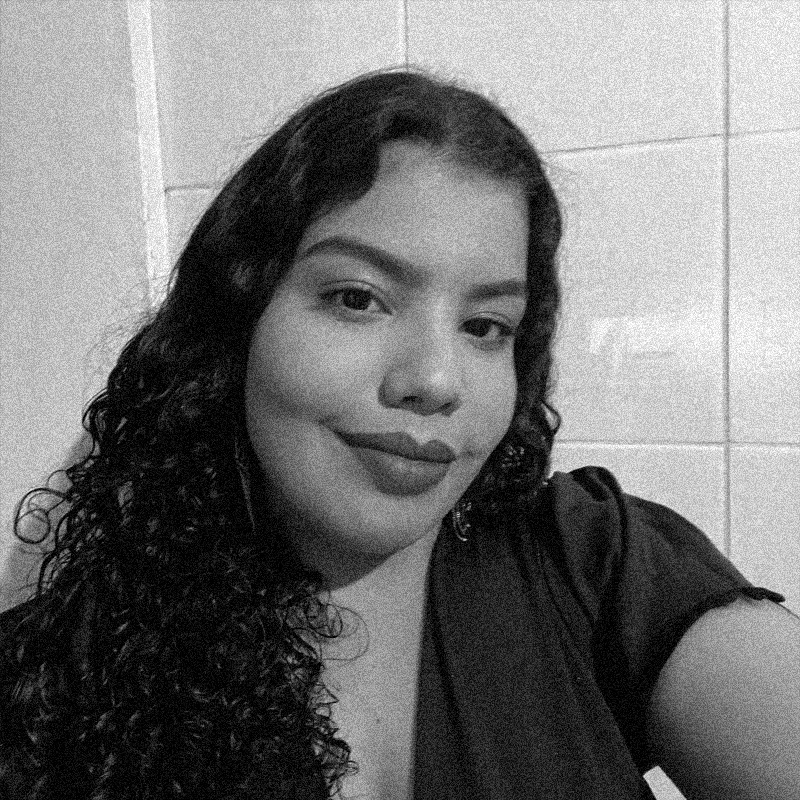 Retrato fotográfico em preto e branco de uma mulher jovem, apresentado dentro de uma moldura circular. Ela tem cabelos escuros, longos e bem cacheados que caem sobre um dos ombros. Ela olha para a câmera com uma expressão serena e um leve sorriso. Veste uma peça de roupa escura e o fundo do retrato mostra uma parede revestida com azulejos claros e retangulares. A imagem mantém a estética de textura granulada e alto contraste, semelhante a uma impressão em papel jornal ou estêncil, comum aos outros retratos da série. O círculo está centralizado sobre um fundo branco com texturas sutis.