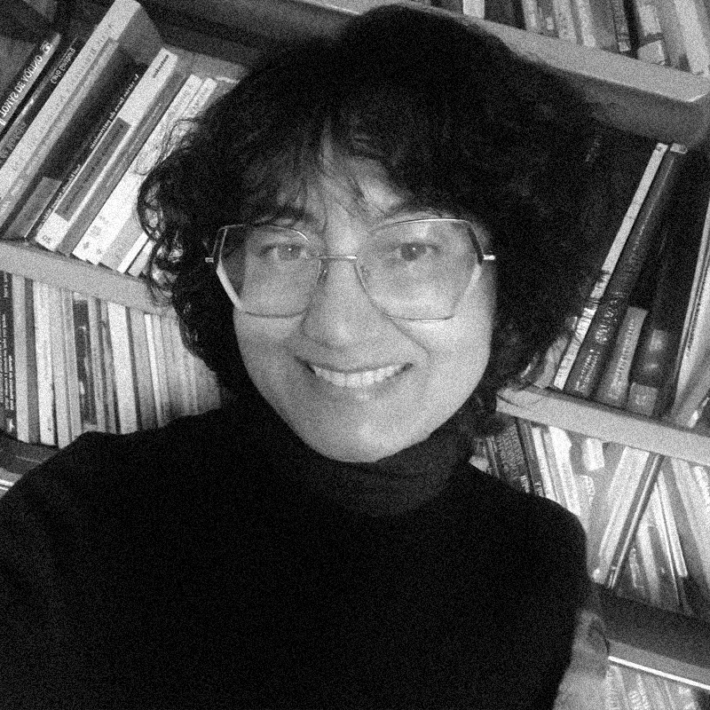 Retrato fotográfico em preto e branco de uma mulher, apresentado dentro de uma moldura circular. Ela sorri para a câmera, tem cabelos escuros, curtos e volumosos, e usa óculos de grau com armação grande e geométrica. Veste uma blusa escura de gola alta. Ao fundo, é possível ver prateleiras de uma estante repletas de livros, sugerindo um ambiente de biblioteca ou escritório. A imagem mantém a estética das anteriores, com uma textura granulada que remete a impressões artísticas ou estêncil. O círculo está centralizado sobre um fundo branco com texturas sutis que lembram papel.