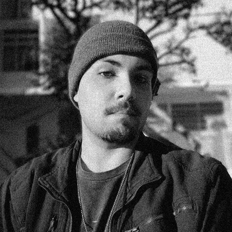 etrato fotográfico em preto e branco de um homem jovem, apresentado dentro de uma moldura circular. Ele tem a pele clara, usa uma touca de lã escura e possui bigode e cavanhaque ralos. Ele olha para a câmera com uma expressão séria. Veste uma jaqueta escura aberta sobre uma camiseta e usa uma corrente no pescoço. Ao fundo, vê-se uma paisagem externa ensolarada com árvores e parte de uma construção. A imagem mantém a estética de textura granulada que remete a impressões artísticas ou estêncil, e o círculo está centralizado sobre um fundo branco texturizado.