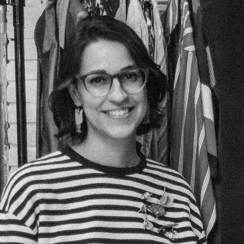 Retrato fotográfico em preto e branco de uma mulher, apresentado dentro de uma moldura circular. Ela sorri para a câmera, tem cabelos escuros curtos e usa óculos de grau com armação escura. Usa brincos médios e retangulares e veste uma blusa com listras horizontais escuras e claras, adornada com um broche em formato de pássaro ou flor. Ao fundo, é possível ver o que parecem ser tecidos ou roupas penduradas. A imagem mantém a estética de textura granulada e alto contraste, semelhante a uma impressão em papel jornal ou técnica de estêncil. O círculo está centralizado sobre um fundo branco com texturas sutis que lembram papel.