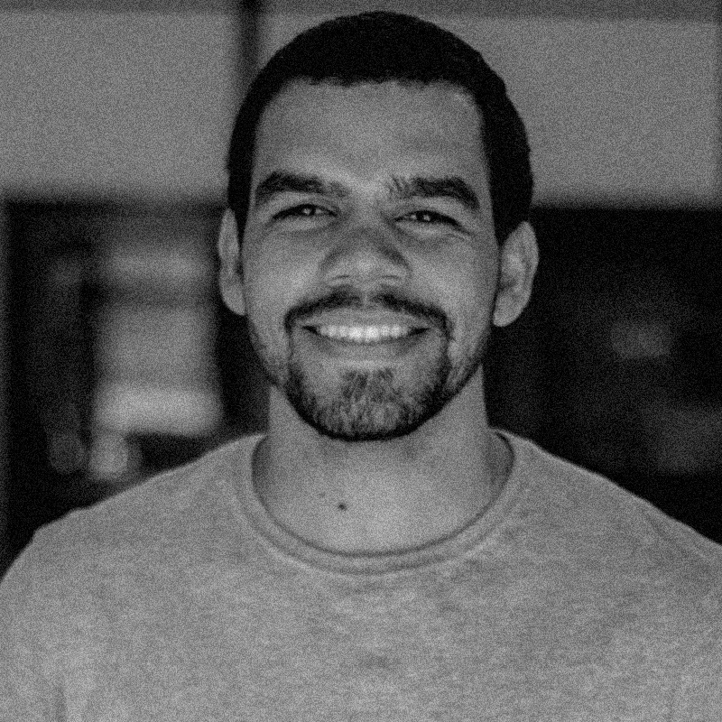 Retrato fotográfico em preto e branco de um homem, apresentado dentro de uma moldura circular. Ele tem cabelos escuros e curtos, e usa óculos de grau com armação escura e arredondada. Ele olha para a câmera com uma expressão tranquila e um leve sorriso. Veste uma blusa escura e o fundo do retrato é liso e claro. A imagem mantém a estética de textura granulada que remete a impressões em papel jornal ou estêncil, seguindo o padrão visual da série. O círculo está centralizado sobre um fundo branco com texturas sutis que lembram papel.