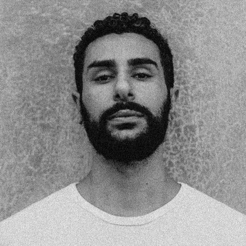 Retrato fotográfico em preto e branco de um homem jovem, apresentado dentro de uma moldura circular. Ele possui a pele morena, cabelos escuros curtos e cacheados, e uma barba e bigode bem aparados e densos. Ele olha diretamente para a câmera com uma expressão séria e calma. Veste uma camiseta branca de gola redonda. O fundo do retrato é uma parede de textura áspera e clara