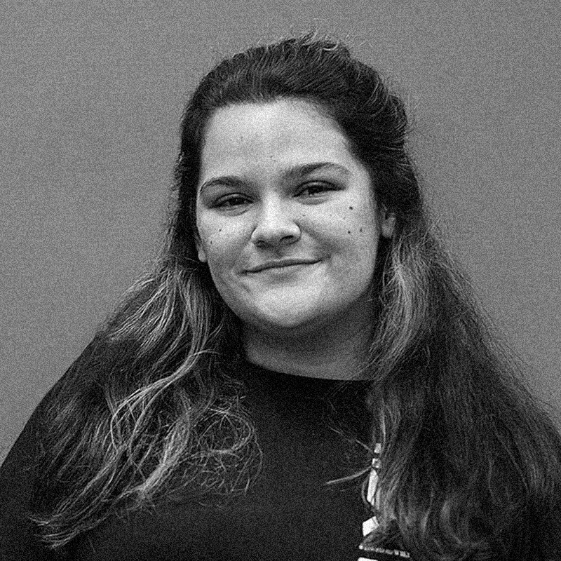 Retrato fotográfico em preto e branco de uma mulher, apresentado dentro de uma moldura circular. Ela tem a pele clara, cabelos escuros e longos que caem sobre os ombros, com as mechas superiores presas para trás. Ela sorri suavemente para a câmera e possui pequenas sardas ou sinais no rosto. Veste uma camiseta escura e o fundo do retrato é liso e neutro. Assim como nos retratos anteriores, a imagem apresenta uma textura granulada característica de impressões artísticas ou estêncil. O círculo está centralizado sobre um fundo branco com texturas sutis que lembram papel.