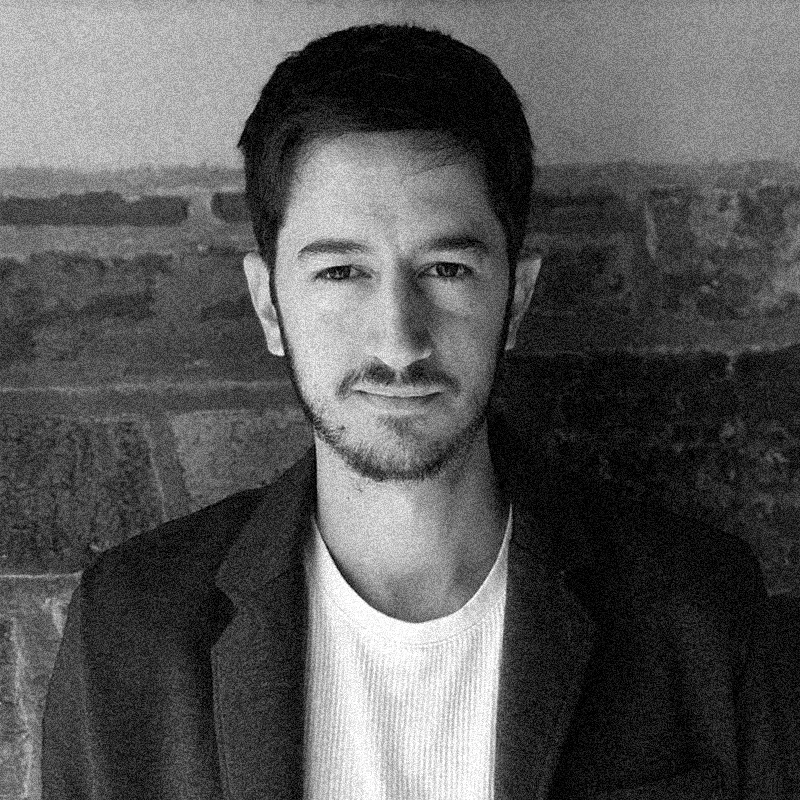 Retrato fotográfico em preto e branco de um homem, apresentado dentro de uma moldura circular. Ele tem a pele clara, cabelos escuros curtos e barba rala. Olha diretamente para a câmera com um leve sorriso. Veste uma camiseta branca de gola redonda sob um paletó ou blazer escuro. O fundo da foto é desfocado e mostra o que parece ser uma paisagem urbana ou arquitetônica. A imagem possui uma textura granulada, seguindo o padrão de impressão artística ou estêncil dos retratos anteriores, e o círculo está centralizado sobre uma base branca texturizada.