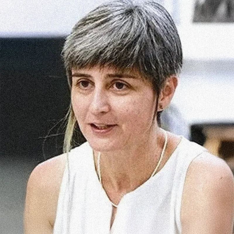 Retrato fotográfico colorido de uma mulher de pele clara. Ela tem cabelos curtos, em um corte estilo "pixie", com fios grisalhos e uma mecha frontal mais longa e descolorida em tom loiro. Ela olha ligeiramente para o lado com uma expressão atenta, como se estivesse em meio a uma conversa. Veste uma blusa branca sem mangas e usa um colar de contas brancas finas. O fundo da imagem está desfocado, sugerindo um ambiente interno iluminado.