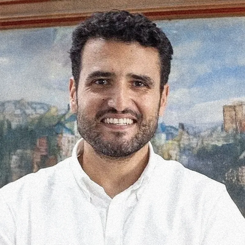 Retrato fotográfico colorido de um homem de pele clara, com cabelos escuros e curtos e barba bem aparada. Ele olha diretamente para a câmera com um sorriso aberto e expressivo.

Veste uma camisa social branca de gola aberta. O fundo da imagem mostra parte de uma pintura ou quadro com moldura de madeira, retratando o que parece ser uma paisagem urbana ou campestre em tons de azul, verde e terra. Assim como em alguns retratos anteriores, a imagem apresenta uma leve textura granulada, mas sem a moldura circular.